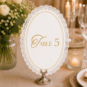 以下が含まれることがあります： 中央に金色の「Table 5」の文字が記された楕円形のテーブルナンバーサイン。二重の金色の縁取りと繊細な白いレースの縁が特徴で、小さな円形の銀色の金属製ペデスタルベースに取り付けられています。