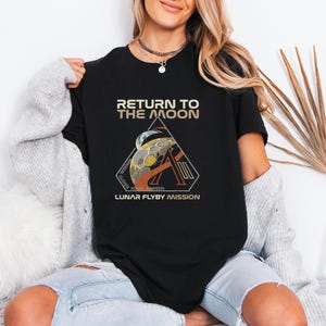 May include: Black t-shirt with a graphic design featuring the text "RETURN TO THE MOON" above a triangular emblem. The emblem includes a stylized moon and the words "LUNAR FLYBY MISSION".