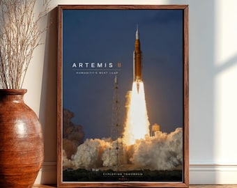May include: A framed poster featuring the Artemis II rocket launching into space. The image shows the rocket with a bright orange and white flame, with the text "ARTEMIS II HUMANITY'S NEXT LEAP EXPLORING TOMORROW NASA". The poster is in a wooden frame.