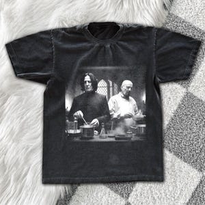 May include: Black t-shirt featuring a grayscale image of two figures in a lab setting. One figure is stirring a pot, while the other stands behind. The shirt has a vintage, washed appearance.