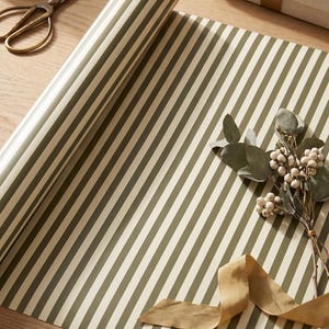 May include: A roll of wrapping paper featuring a repeating pattern of vertical olive green and cream stripes. The paper is partially unrolled, displaying the striped design across its surface, accompanied by a sprig of dried eucalyptus and ribbon.