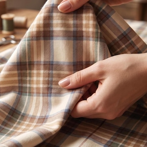 May include: A woven fabric featuring a classic plaid pattern in shades of beige, cream, brown, and muted blue. The textile displays a structured grid design with intersecting horizontal and vertical lines of varying widths, creating a balanced tartan aesthetic.