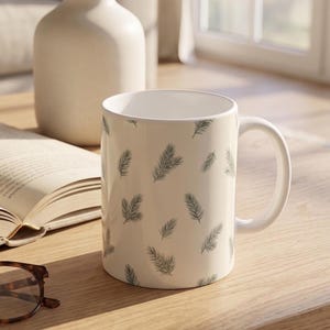 May include: A white ceramic mug featuring a repeating pattern of small, dark green pine needle sprigs. The mug has a smooth, cylindrical body with a curved handle on the right side and a clean, white interior finish, standing upright on a flat surface.