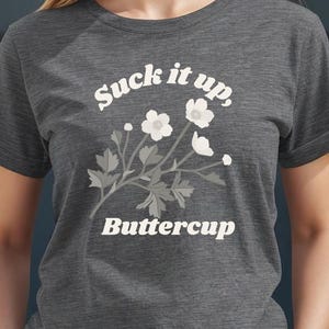 May include: A gray t-shirt with the text "Suck it up, Buttercup" in white, along with a floral design. The shirt is short-sleeved and made of a soft material. The model is wearing blue jeans.