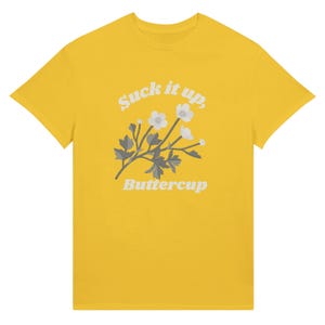 May include: A yellow t-shirt featuring the text "Suck it up, Buttercup" in a white, stylized font. Below the text is a floral design with white flowers and gray leaves. The t-shirt is a classic crew neck style.