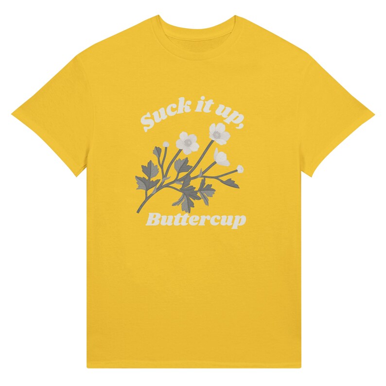 May include: A yellow t-shirt featuring the text "Suck it up, Buttercup" in a white, stylized font. Below the text is a floral design with white flowers and gray leaves. The t-shirt is a classic crew neck style.