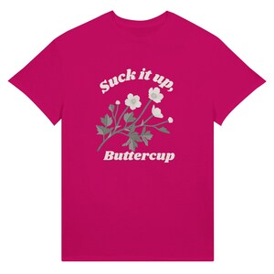 May include: A magenta t-shirt featuring the text "Suck it up, Buttercup" in white script above a floral illustration. The design includes white flowers and green leaves.