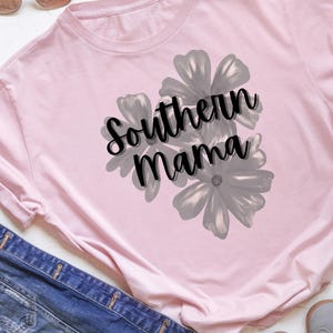 May include: A light pink long-sleeved shirt with the text "Southern Mama" in black script, overlaid on a gray floral design. Sunglasses and denim jeans are visible in the background.