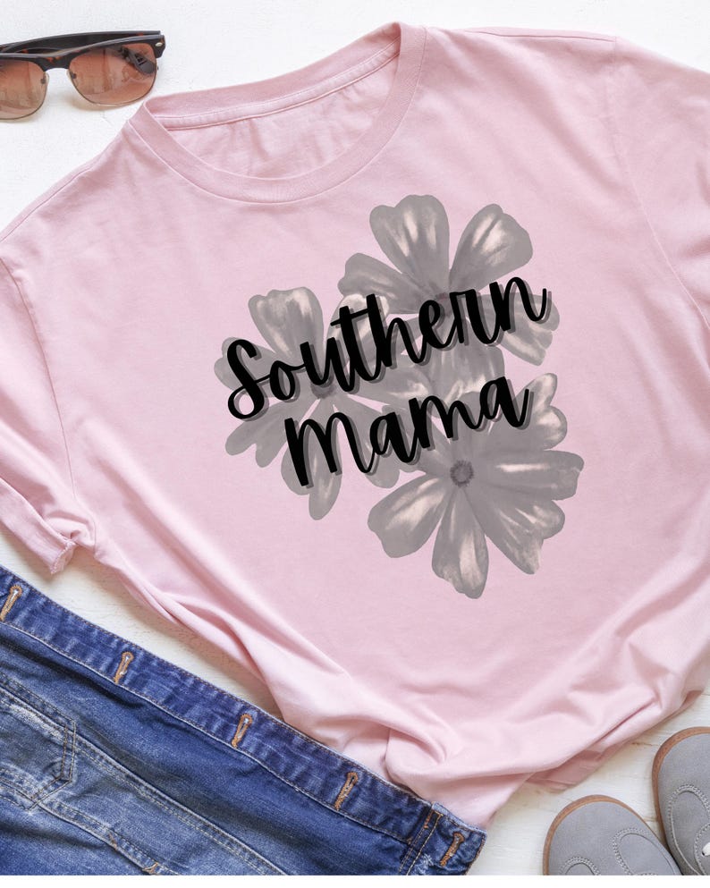 May include: A light pink long-sleeved shirt with the text "Southern Mama" in black script, overlaid on a gray floral design. Sunglasses and denim jeans are visible in the background.