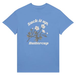 May include: A light blue t-shirt featuring the text "Suck it up, Buttercup" in a whimsical font, accompanied by a floral illustration. The design includes white flowers and gray leaves. The shirt is a classic crew neck style.