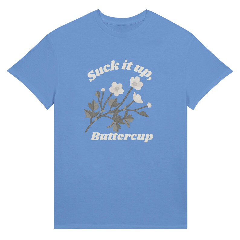 May include: A light blue t-shirt featuring the text "Suck it up, Buttercup" in a whimsical font, accompanied by a floral illustration. The design includes white flowers and gray leaves. The shirt is a classic crew neck style.
