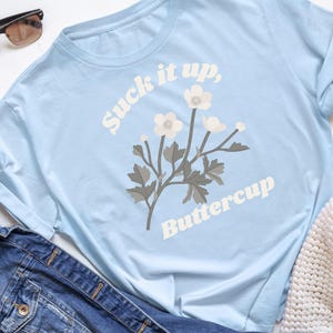 May include: Light blue long-sleeve t-shirt with the text "Suck it up, Buttercup" and a floral graphic. The shirt is made of a soft material and is paired with a denim jacket and sunglasses.