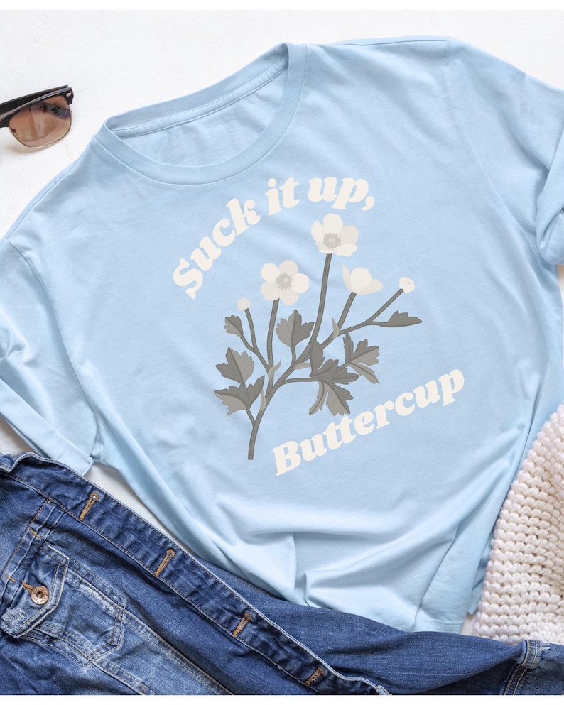 May include: Light blue long-sleeve t-shirt with the text "Suck it up, Buttercup" and a floral graphic. The shirt is made of a soft material and is paired with a denim jacket and sunglasses.