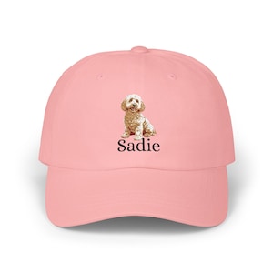 May include: A light pink baseball cap featuring a centered graphic of a tan-colored dog sitting down. Below the dog illustration, the name Sadie is printed in a simple, black, serif font. The cap has a structured crown and a curved brim.