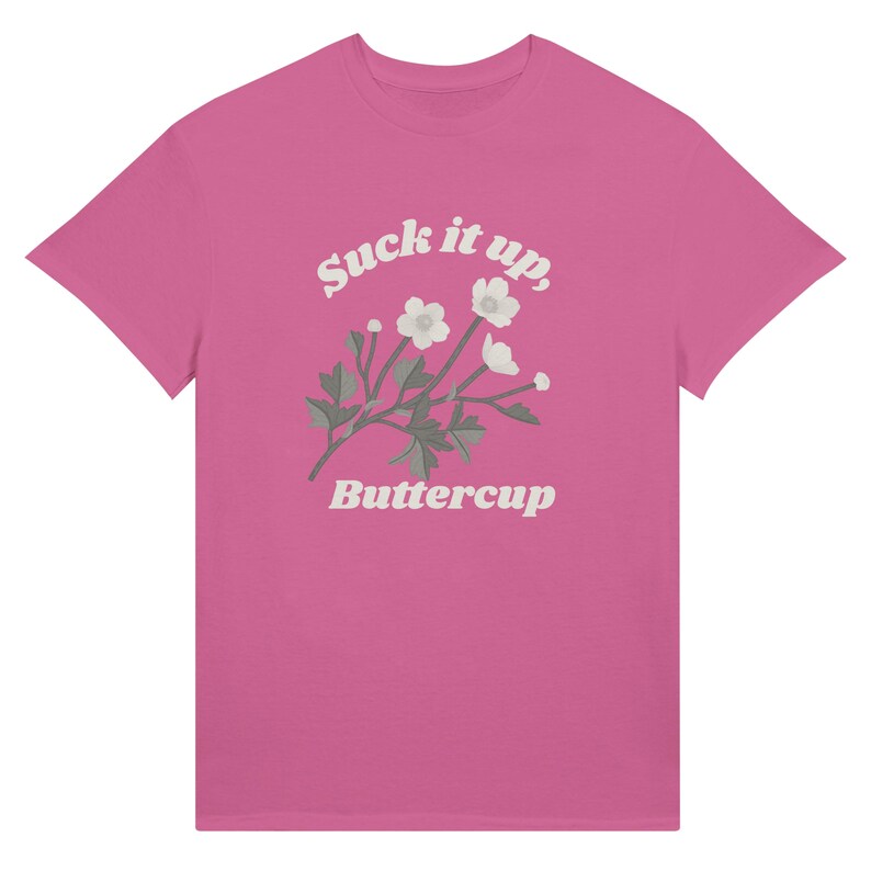 May include: A pink t-shirt featuring the text "Suck it up, Buttercup" in a stylized font, along with a floral illustration of white flowers and green leaves. The shirt is a solid pink color.