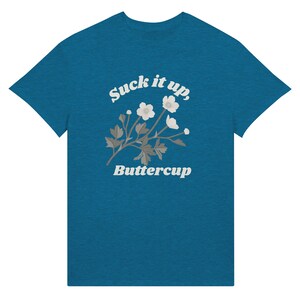 May include: Teal t-shirt with the text "Suck it up, Buttercup" in white script above a floral design. The floral design features white flowers and gray leaves. The t-shirt is a solid teal color.