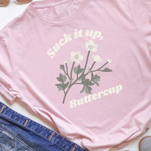 May include: A light pink t-shirt with the text "Suck it up, Buttercup" in white, along with a floral graphic. The shirt is paired with blue denim and sunglasses.