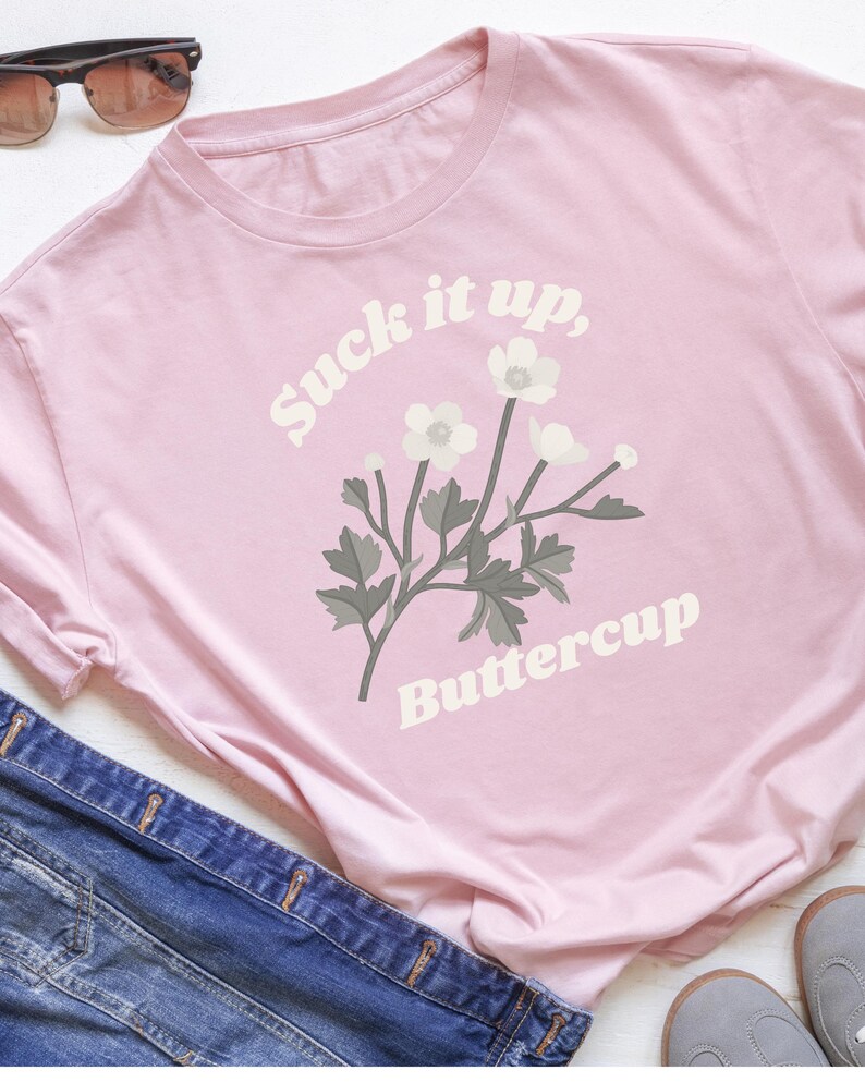 May include: A light pink t-shirt with the text "Suck it up, Buttercup" in white, along with a floral graphic. The shirt is paired with blue denim and sunglasses.