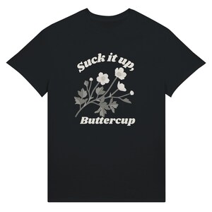 May include: Black t-shirt featuring the text "Suck it up, Buttercup" in a white, stylized font. A floral design with white flowers and gray leaves is centered below the text. The shirt is a classic crew neck style.