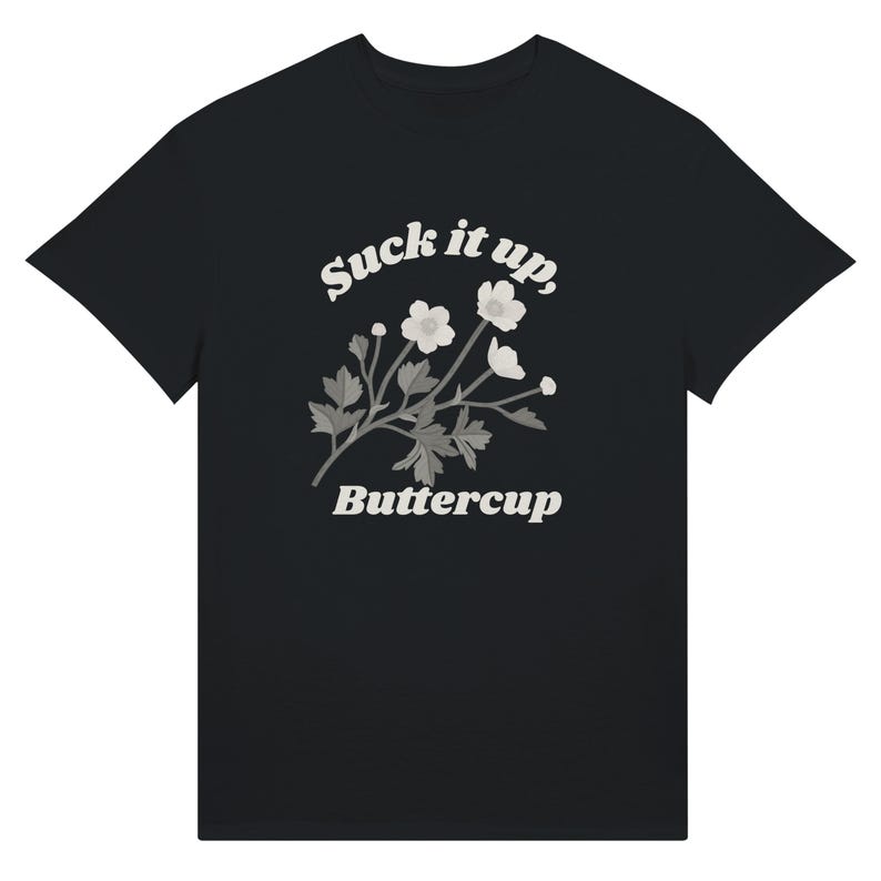 May include: Black t-shirt featuring the text "Suck it up, Buttercup" in a white, stylized font. A floral design with white flowers and gray leaves is centered below the text. The shirt is a classic crew neck style.
