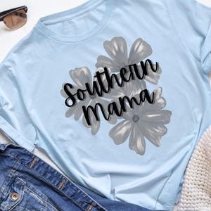 May include: Light blue long-sleeve shirt with the text "Southern Mama" in black script over a gray floral design. Sunglasses and a denim jacket are visible in the background.