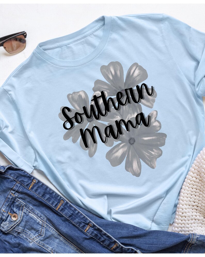 May include: Light blue long-sleeve shirt with the text "Southern Mama" in black script over a gray floral design. Sunglasses and a denim jacket are visible in the background.