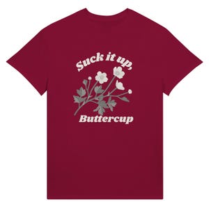 May include: A maroon t-shirt featuring the text "Suck it up, Buttercup" in a white, stylized font. A floral illustration with white flowers and gray leaves is centered below the text. The t-shirt is made of a soft material.
