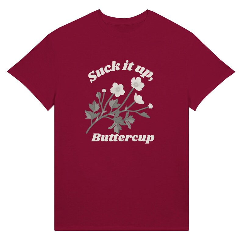 May include: A maroon t-shirt featuring the text "Suck it up, Buttercup" in a white, stylized font. A floral illustration with white flowers and gray leaves is centered below the text. The t-shirt is made of a soft material.