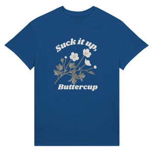 May include: A blue t-shirt featuring the text "Suck it up, Buttercup" in a white, stylized font. Below the text is a floral design with white flowers and gray leaves. The t-shirt is a solid blue color.