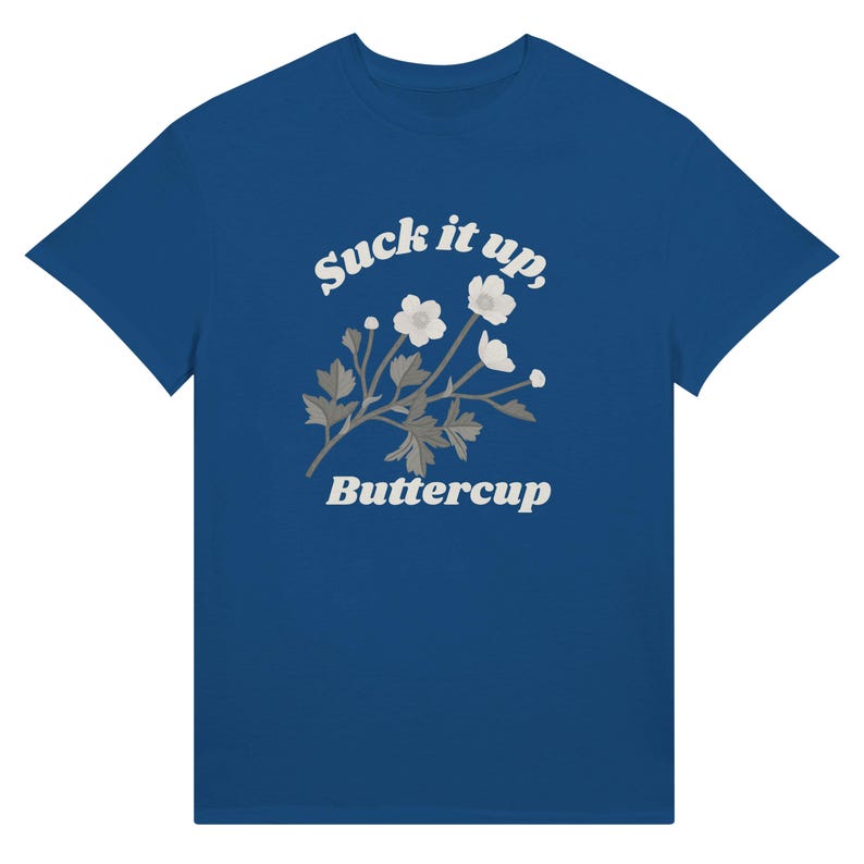 May include: A blue t-shirt featuring the text "Suck it up, Buttercup" in a white, stylized font. Below the text is a floral design with white flowers and gray leaves. The t-shirt is a solid blue color.