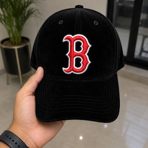 Puede incluir: Una gorra de béisbol negra con una letra B bordada en rojo con un borde blanco en el panel frontal. La gorra tiene una corona estructurada, una visera curva y pequeños ojales de ventilación en la parte superior de los paneles de tela.