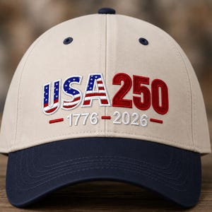 Op de afbeelding: Een beige honkbalpet met een donkerblauwe klep en knoop aan de bovenkant. De voorkant is voorzien van geborduurde tekst USA 250, waarbij USA is gevuld met een Amerikaanse vlagpatroon en 250 in het rood. Daaronder staat de witte tekst 1776-2026.