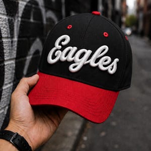 Könnte beinhalten: Eine schwarze Baseballkappe mit rotem Schirm und roten Ösen. Das Wort Eagles ist in weißer, erhabener Schreibschrift auf die Vorderseite gestickt. Die Kappe verfügt über eine strukturierte Krone und einen gebogenen Schirm mit Kontrast.