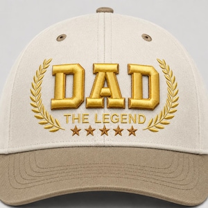 Puede incluir: Una gorra de béisbol beige con visera marrón y ojales marrones. El frente presenta un texto bordado en dorado que dice DAD THE LEGEND, flanqueado por dos ramas de laurel doradas y cinco estrellas doradas en una fila horizontal debajo.