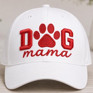 Puede incluir: Una gorra de béisbol blanca con texto bordado en rojo que dice DOG, con una huella de pata roja reemplazando la letra O, y la palabra mama bordada en letra cursiva roja debajo del diseño de la huella en el panel frontal.