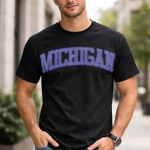 May include: Black t-shirt with the word "MICHIGAN" in a blue, collegiate-style font across the chest. The shirt is short-sleeved and worn by a person wearing blue jeans and a watch.