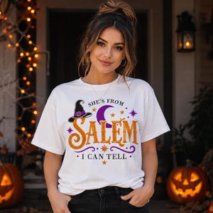 Puede incluir: Una camiseta blanca de manga corta con un diseño gráfico que incluye el texto "SHE'S FROM SALEM I CAN TELL" en naranja y negro. El diseño presenta una luna creciente morada, un sombrero de bruja negro y estrellas naranjas sobre fondo blanco.