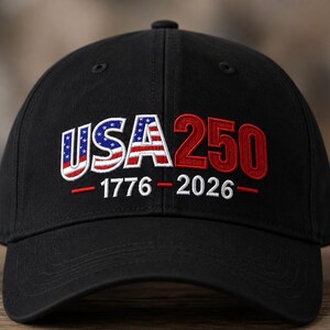 Op de afbeelding: Een zwarte baseballpet met geborduurde tekst op de voorzijde. De letters USA zijn gevuld met een rood, wit en blauw Amerikaans vlagpatroon, gevolgd door het getal 250 in het rood. Daaronder is de tekst 1776 - 2026 in het wit geborduurd.