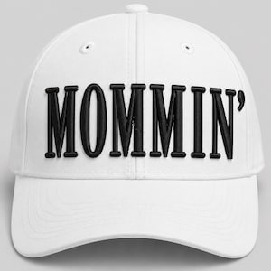 Puede incluir: Gorra de béisbol blanca con la palabra "MOMMIN'" bordada en letras negras en la parte delantera. La gorra tiene una visera curva y un botón en la parte superior. Un accesorio casual.