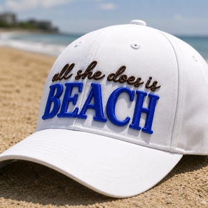 Puede incluir: Una gorra de béisbol blanca con la frase bordada "all she does is BEACH" en marrón y azul. La gorra está sobre una playa de arena con el océano al fondo.