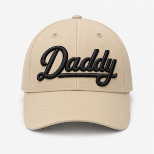 Op de afbeelding: Een beige honkbalpet met het woord Daddy geborduurd in verhoogde zwarte schrijfletters op de voorpanelen. De pet heeft een gestructureerde kroon, een gebogen klep en bijpassende ton-sur-ton stiksels over de gehele constructie.
