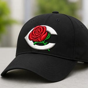 Puede incluir: Una gorra de béisbol negra con un diseño bordado en la parte frontal. El diseño consiste en una letra C grande de color blanco con una rosa roja detallada, un tallo y hojas verdes situados dentro de la curva. La gorra tiene una visera curva.