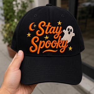 Könnte beinhalten: Eine schwarze Baseballkappe mit orangefarbenem Sticktext, der Stay Spooky lautet. Das Design umfasst ein kleines weißes Gespenst, eine Mondsichel und mehrere gelbe Sterne, die auf die Vorderseite der Kappe über dem Schirm gestickt sind.