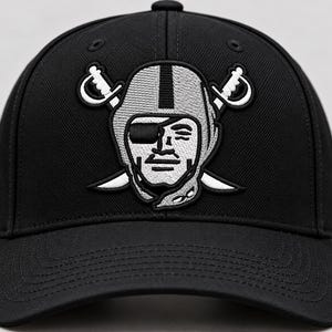 Puede incluir: Una gorra de béisbol negra con un logotipo bordado de una persona con casco de fútbol americano y un parche en el ojo, situada frente a dos espadas cruzadas. El diseño está realizado con hilo plateado, blanco y negro en el panel frontal.