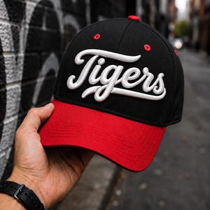 May include: A black baseball cap with a red brim and a red button on top. The word Tigers is embroidered in white script across the front panels. The cap features red eyelets and structured stitching on the brim, creating a bold contrast.