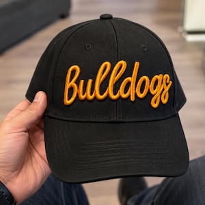 Puede incluir: Una gorra de béisbol negra con la palabra "bulldogs" bordada en relieve con una tipografía cursiva de color amarillo dorado en los paneles frontales. La gorra tiene una corona estructurada, visera curva con costuras y un botón superior.