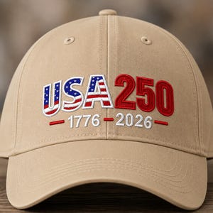 Op de afbeelding: Een beige honkbalpet met geborduurde tekst op de voorkant. De letters USA zijn gevuld met een rood, wit en blauw sterren- en strepenpatroon. Het getal 250 is in het rood. Daaronder staat 1776 - 2026 geborduurd in het wit met rode streepjes.
