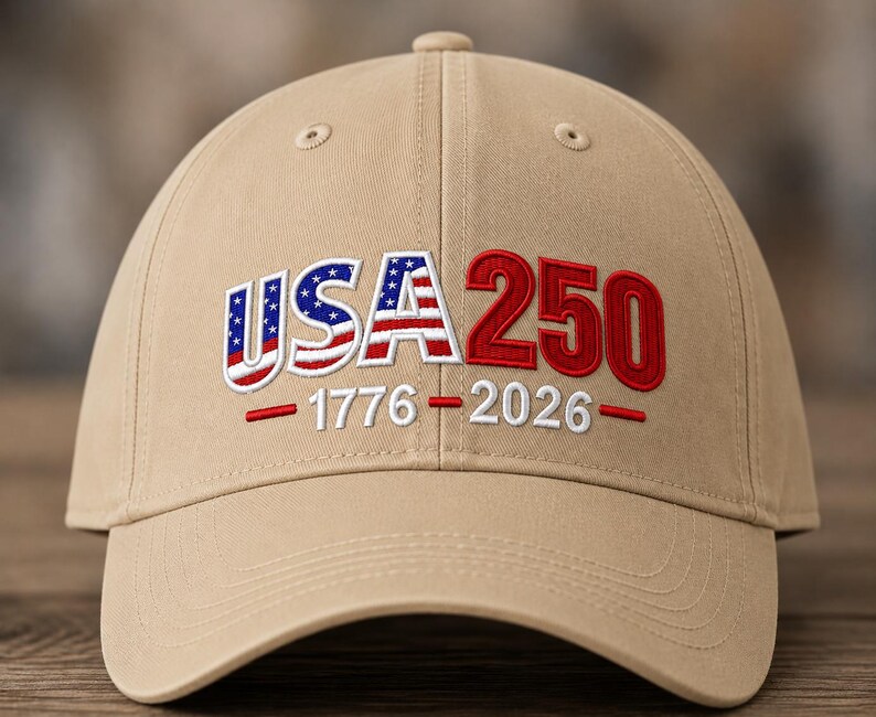 Op de afbeelding: Een beige honkbalpet met geborduurde tekst op de voorkant. De letters USA zijn gevuld met een rood, wit en blauw sterren- en strepenpatroon. Het getal 250 is in het rood. Daaronder staat 1776 - 2026 geborduurd in het wit met rode streepjes.