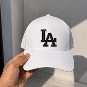 May include: A white baseball cap featuring a black embroidered LA logo on the front. The cap has a structured crown, a curved brim, and stitched eyelets on the top panels. The design is clean and simple, focusing on the contrast between the white fabric and the bold black lettering.