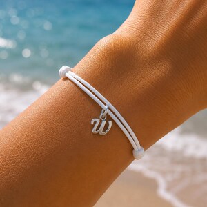 Op de afbeelding: Een wit koordarmbandje met een zilverkleurige metalen bedel in de vorm van de kleine letter w. De armband is gemaakt met een dubbel wit koordontwerp en bevat twee kleine witte kralen aan de uiteinden van het koord.
